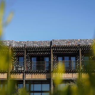 Stone building with a wooden pergola and balcony, clear blue sky behind, and greenery in the foreground with a white umbrella at the bottom blends in.