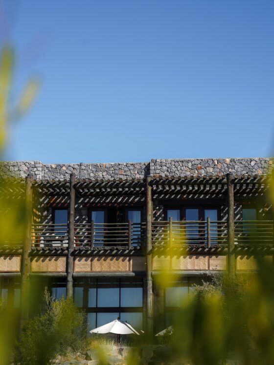 Stone building with a wooden pergola and balcony, clear blue sky behind, and greenery in the foreground with a white umbrella at the bottom blends in.