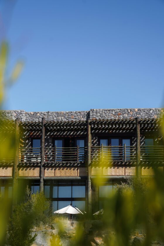 Stone building with a wooden pergola and balcony, clear blue sky behind, and greenery in the foreground with a white umbrella at the bottom blends in.