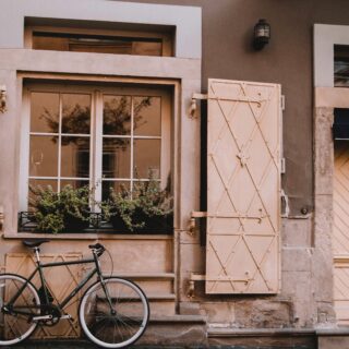 Exterior of a beige stone hotel facade with pale decorative shutters and a bicycle leaning against the wall beneath a window.