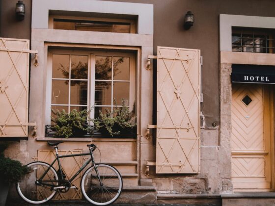 Exterior of a beige stone hotel facade with pale decorative shutters and a bicycle leaning against the wall beneath a window.