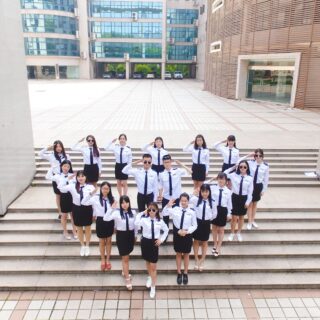 Group of students in white uniform shirts and navy skirts/pants stand on outdoor steps, saluting toward the camera in a campus courtyard.