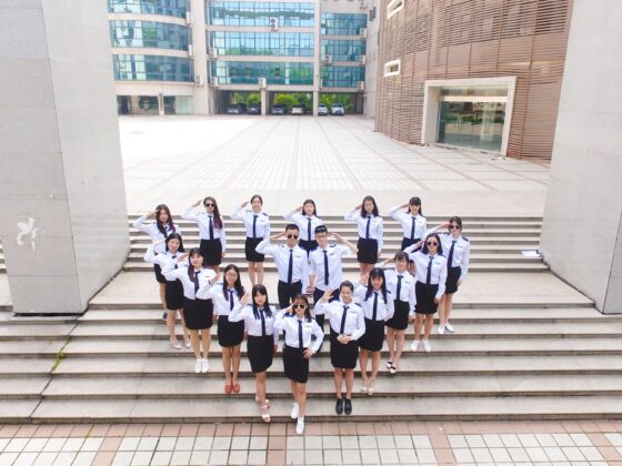 Group of students in white uniform shirts and navy skirts/pants stand on outdoor steps, saluting toward the camera in a campus courtyard.