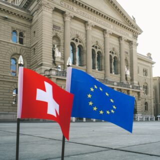 Front of a historic government building with columned façade and arched windows; two flags—Swiss (red with white cross) and European Union—fly in the foreground.