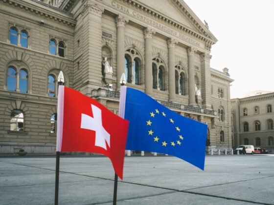 Front of a historic government building with columned façade and arched windows; two flags—Swiss (red with white cross) and European Union—fly in the foreground.