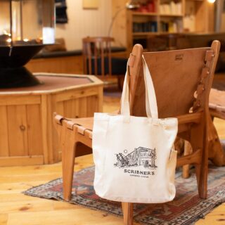 Canvas tote bag hanging on a wooden chair in a cozy lodge lobby with a stone fireplace and bookshelves in the background.