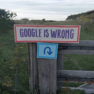 Sign reading 'GOOGLE IS WRONG' tacked to a weathered wooden fence in a rural field.
