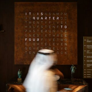 Motion-blurred person in white walks past a large rust-colored panel with a grid of letters.
