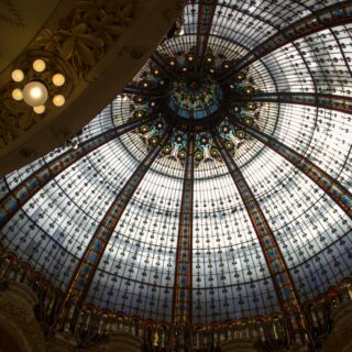 Interior view of a grand glass-domed ceiling with ornate stained-glass panels and a central chandelier.