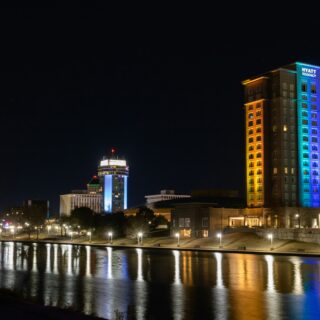 Night view of a riverside city with a tall Hyatt Regency hotel lit in rainbow colors.