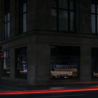 Nighttime street corner with a dark stone building and large windows; red light trails sweep along the street below.