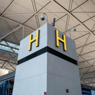 Interior of a modern airport terminal with a gray pillar bearing yellow 'H' signs behind security cameras at the top.