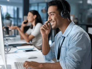 Support agent wearing a headset at a computer in a bright open-plan office.