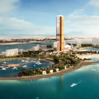 A futuristic coastal development with a circular marina of yachts, a wraparound breakwater, and a tall reddish skyscraper on a tree-filled peninsula under a blue sky