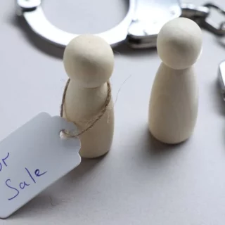 Two wooden peg dolls with a 'For Sale' tag, beside a pair of metal handcuffs in the background.