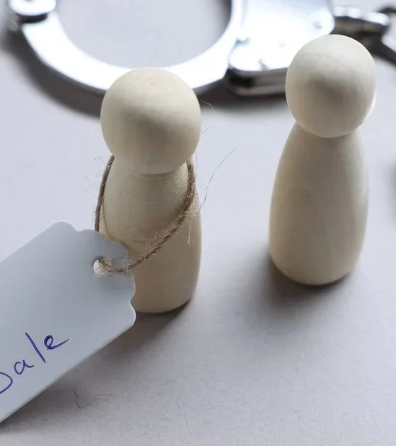 Two wooden peg dolls with a 'For Sale' tag, beside a pair of metal handcuffs in the background.