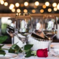 Wine glasses on a dining table with a red rose and green leaves, warm bokeh lights in the background, suggesting a romantic dinner setting.