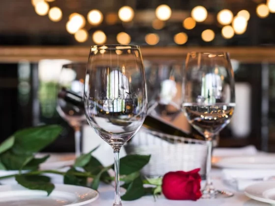 Wine glasses on a dining table with a red rose and green leaves, warm bokeh lights in the background, suggesting a romantic dinner setting.