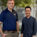Two men stand side by side outdoors, smiling at the camera; one wears a navy polo shirt and the other a dark blue button-down shirt, with trees and a modern building in the background.