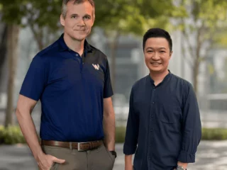 Two men stand side by side outdoors, smiling at the camera; one wears a navy polo shirt and the other a dark blue button-down shirt, with trees and a modern building in the background.