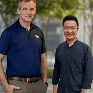 Two men stand side by side outdoors, smiling at the camera; one wears a navy polo shirt and the other a dark blue button-down shirt, with trees and a modern building in the background.