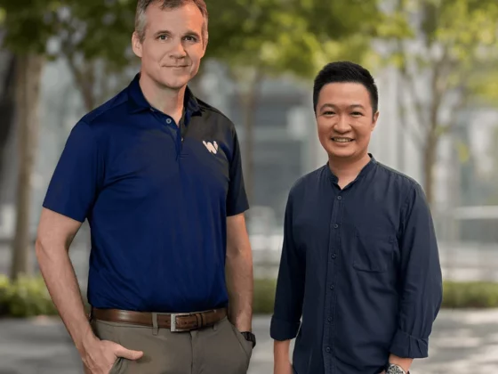 Two men stand side by side outdoors, smiling at the camera; one wears a navy polo shirt and the other a dark blue button-down shirt, with trees and a modern building in the background.
