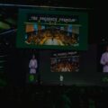 Speaker on a dark stage at a conference, large screens show 'The Presence Premium' and a crowded audience.
