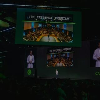 Speaker on a dark stage at a conference, large screens show 'The Presence Premium' and a crowded audience.