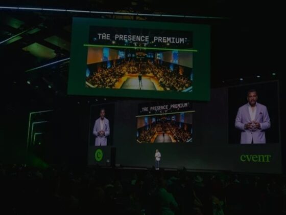 Speaker on a dark stage at a conference, large screens show 'The Presence Premium' and a crowded audience.
