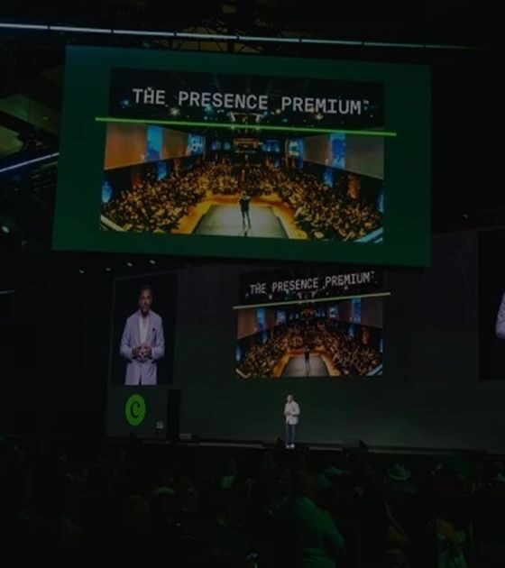 Speaker on a dark stage at a conference, large screens show 'The Presence Premium' and a crowded audience.