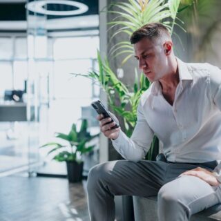 Man in a white shirt and gray pants sits on a bench in a bright office lobby, looking at his smartphone.