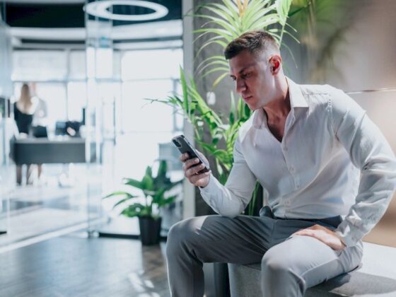 Man in a white shirt and gray pants sits on a bench in a bright office lobby, looking at his smartphone.