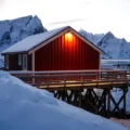 Red wooden cabin on stilts over a frozen inlet, warm light at the door, surrounded by snow-capped mountains in winter.