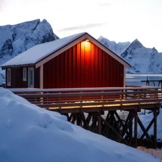 Red wooden cabin on stilts over a frozen inlet, warm light at the door, surrounded by snow-capped mountains in winter.