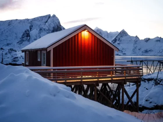 Red wooden cabin on stilts over a frozen inlet, warm light at the door, surrounded by snow-capped mountains in winter.
