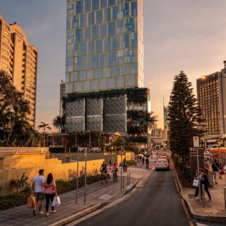 City street at sunset with pedestrians, cyclists, and a tram stop beside modern glass buildings.