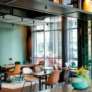 A stylish, modern cafe interior with teal-green tiled columns, mixed chairs, and large windows letting in daylight.