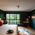 Hotel room interior with a large bed, red accent pillows, dark green walls, and a window view; chair, desk, and floor lamp nearby.