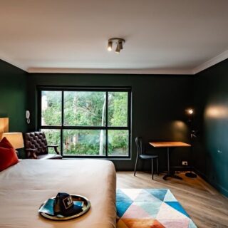 Hotel room interior with a large bed, red accent pillows, dark green walls, and a window view; chair, desk, and floor lamp nearby.