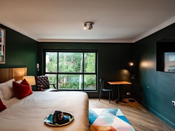Hotel room interior with a large bed, red accent pillows, dark green walls, and a window view; chair, desk, and floor lamp nearby.