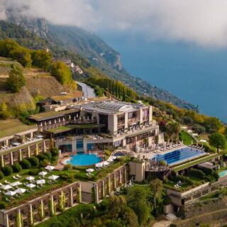 Luxury hillside resort with multiple terraces, a central round pool, and sun loungers overlooking the blue sea below.