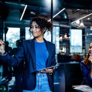 Woman in a blue blazer writes on a glass wall with a marker, holding a tablet, as a colleague watches in a modern office conference room.