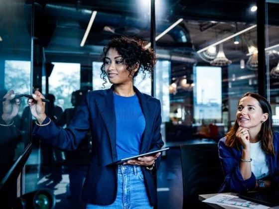 Woman in a blue blazer writes on a glass wall with a marker, holding a tablet, as a colleague watches in a modern office conference room.