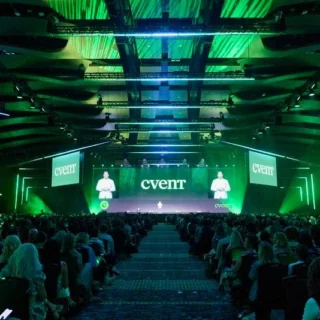 Crowd seated in a large conference hall facing a neon-green stage; large screen reads 'Cvent' with presenters on either side.