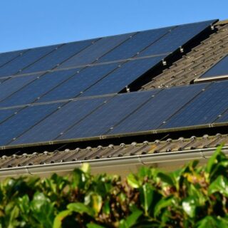 Solar panels installed on a sloped roof with a blue sky and green hedge in the foreground.