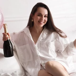 Woman sitting on a bed holding a champagne bottle and a glass, smiling at a pink balloon party scene at home.