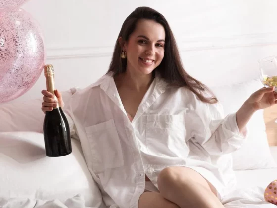 Woman sitting on a bed holding a champagne bottle and a glass, smiling at a pink balloon party scene at home.