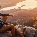 Two people sit on a rocky outcrop at sunset, overlooking a valley with distant water and warm golden light.