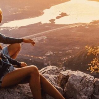 Two people sit on a rocky outcrop at sunset, overlooking a valley with distant water and warm golden light.
