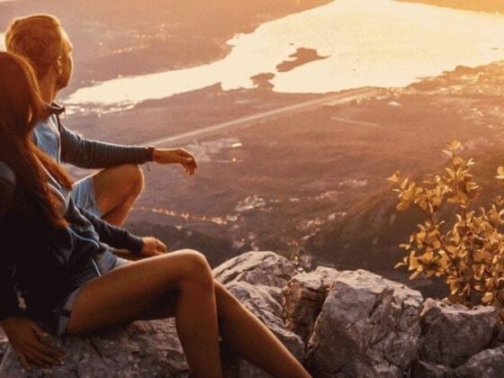 Two people sit on a rocky outcrop at sunset, overlooking a valley with distant water and warm golden light.